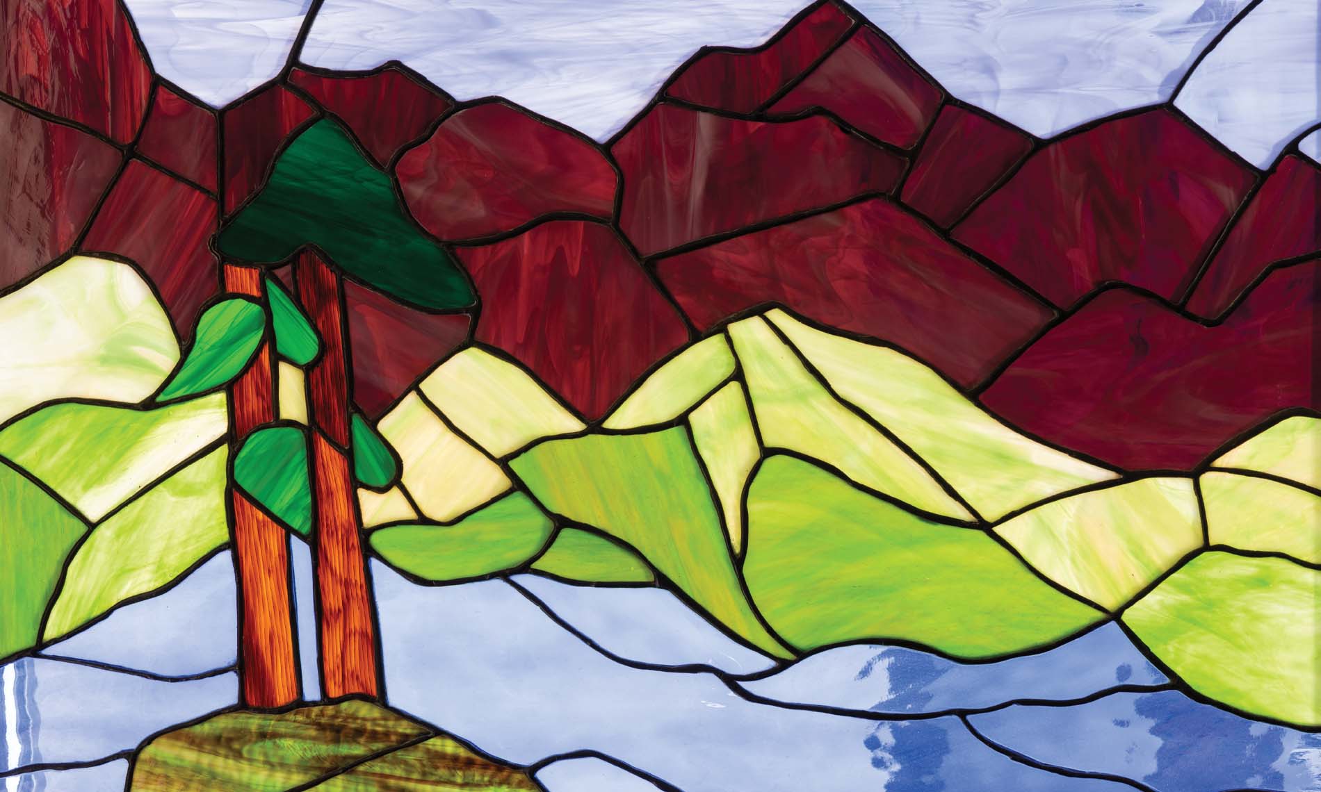 A piece of stained glass art of mountains, fields, and trees.