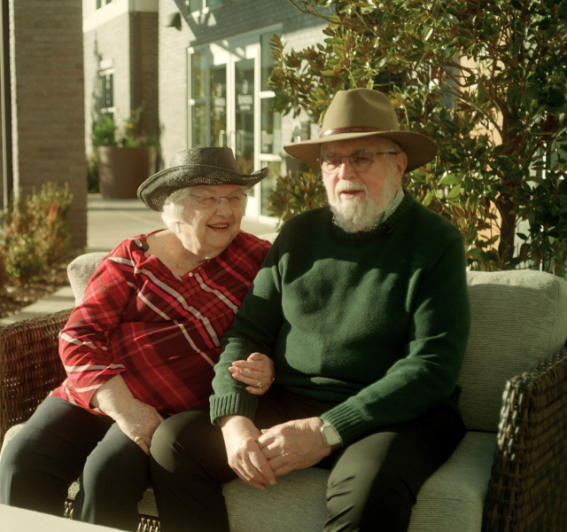 Wife and husband sitting outside and smiling. Her arm is linked into his. 